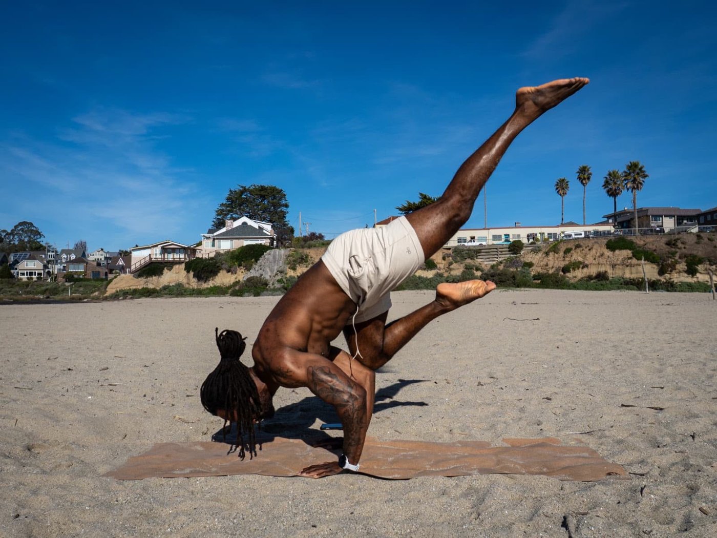 A man holding an arm balance on a mat at the beach, legs extended into the air