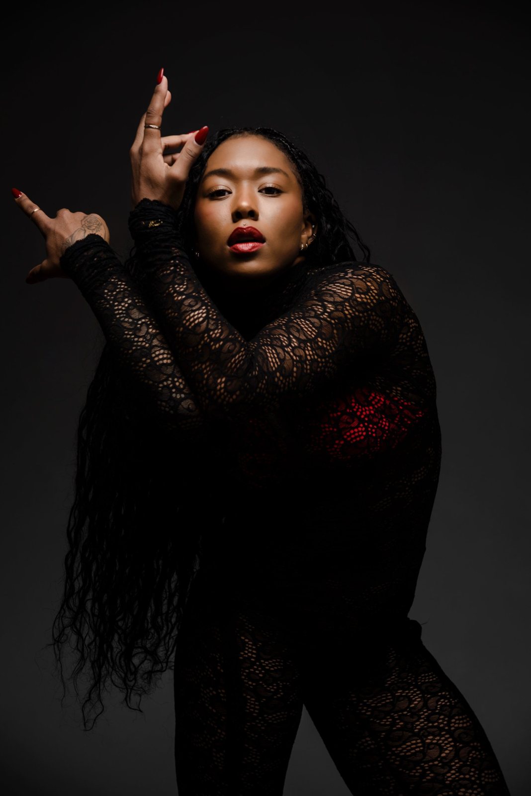 Morgan Simone in a black lace bodysuit, posed with her hands raised against a dark background